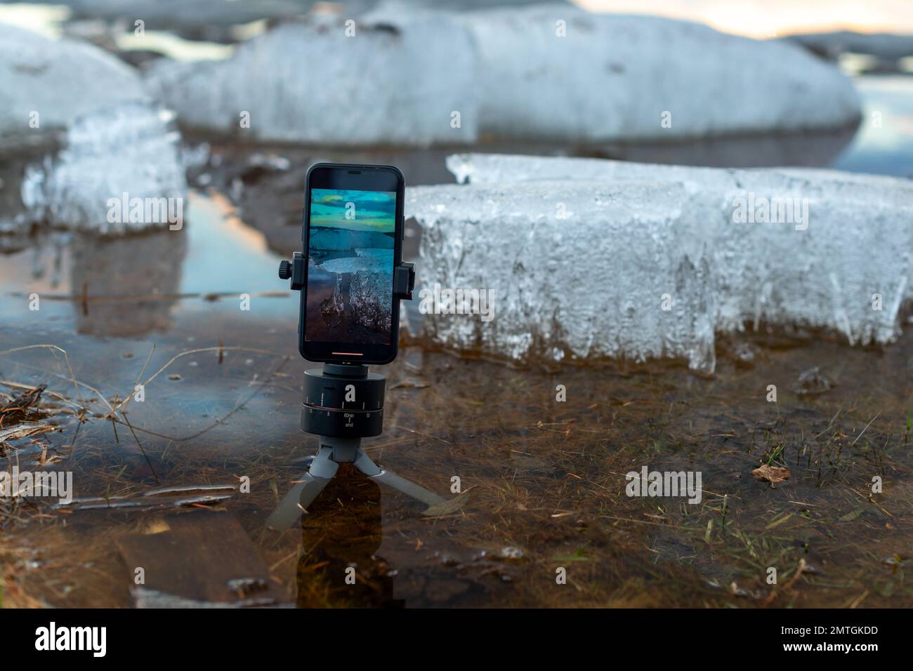 Der Bildschirm eines Mobiltelefons macht ein Foto des Quelleises, der auf einem kleinen Stativ im Fluss steht, vor dem Hintergrund von Eisschollen in der Sonne Stockfoto