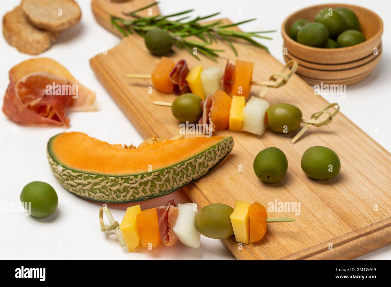 Ein Stück Melone. Käse, Oliven, Jamon auf einem Spieß. Grüne Oliven in einer Holzschüssel. Draufsicht. Weißer Hintergrund. Stockfoto
