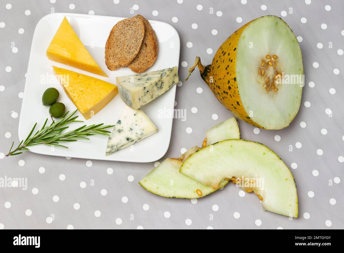 Käse, Brot, grüne Oliven und Rosmarin auf einem weißen Teller. Melonenscheiben auf dem Tisch. Flach verlegt. Grauer weißer Hintergrund mit Punktmuster. Stockfoto