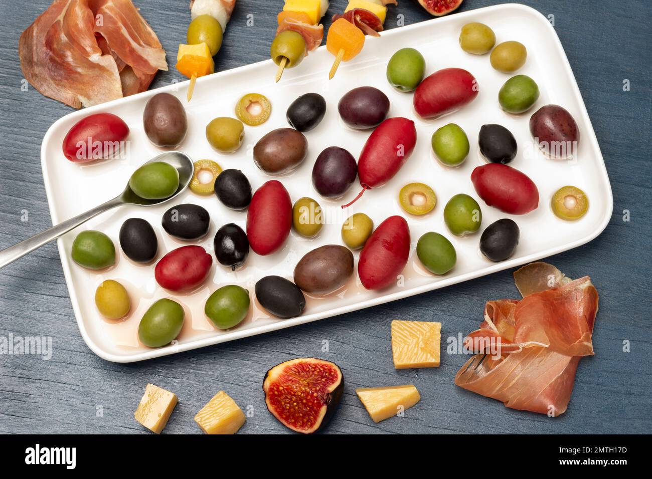 Oliven verschiedener Sorten und Farben als weiße Platte. Käse, Oliven, Jamon und Feigen auf einem Tisch. Flach verlegt. Blauer Hintergrund. Stockfoto