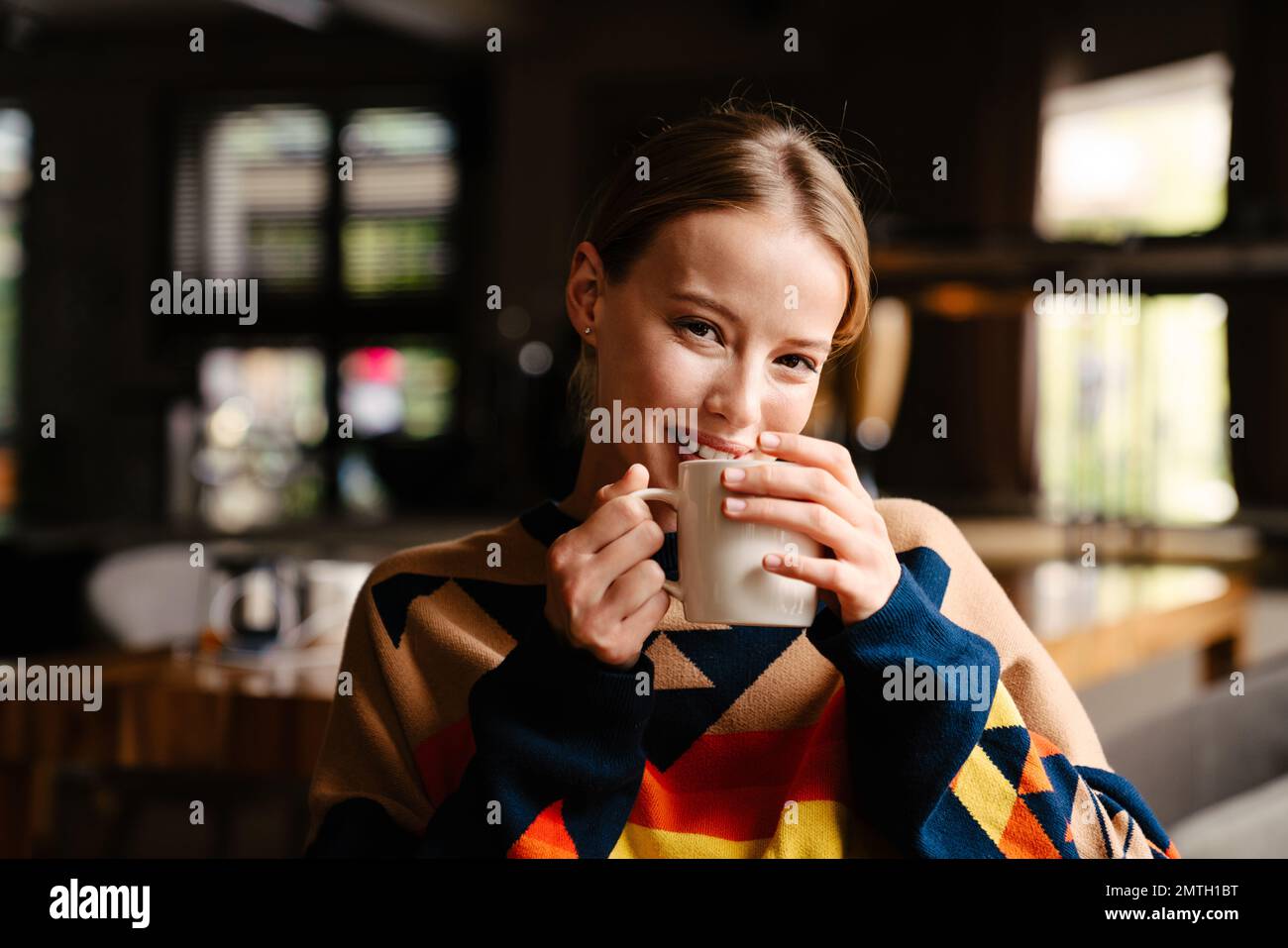 Fröhliche, schöne Frau, die einen farbenfrohen Pullover trägt und Tee trinkt, während sie im Café sitzt Stockfoto