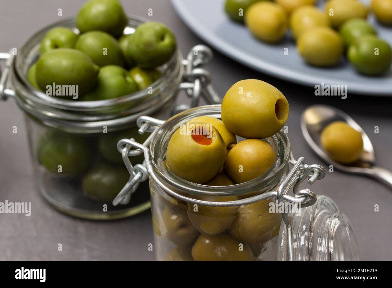 Grüne Oliven in Glasgefäßen. Oliven auf einem blauen Teller. Schließen. Grauer Hintergrund. Stockfoto