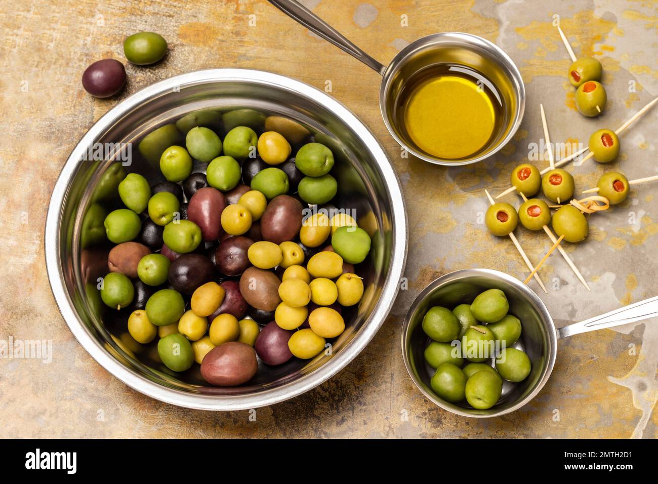 Oliven in einer Metallschüssel. Olivenöl in einer Schüssel. Oliven auf Spießen. Flach verlegt. Rostiger Hintergrund. Stockfoto