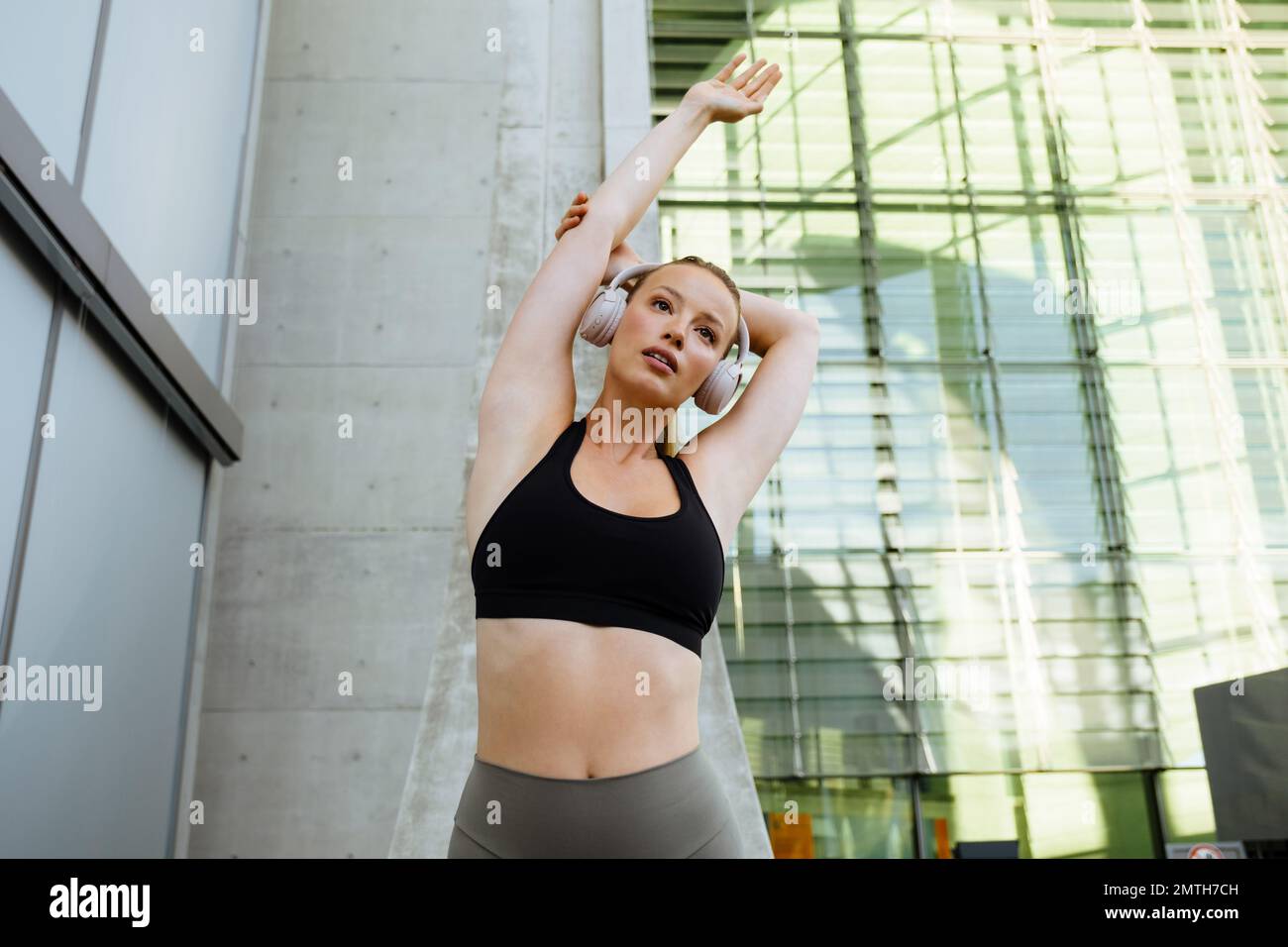 Junge wunderschöne, ruhige Frau mit Kopfhörern und schwarzem Oberteil, die ihren Arm nach außen streckt, während sie in der Nähe eines modernen Gebäudes im Freien steht Stockfoto