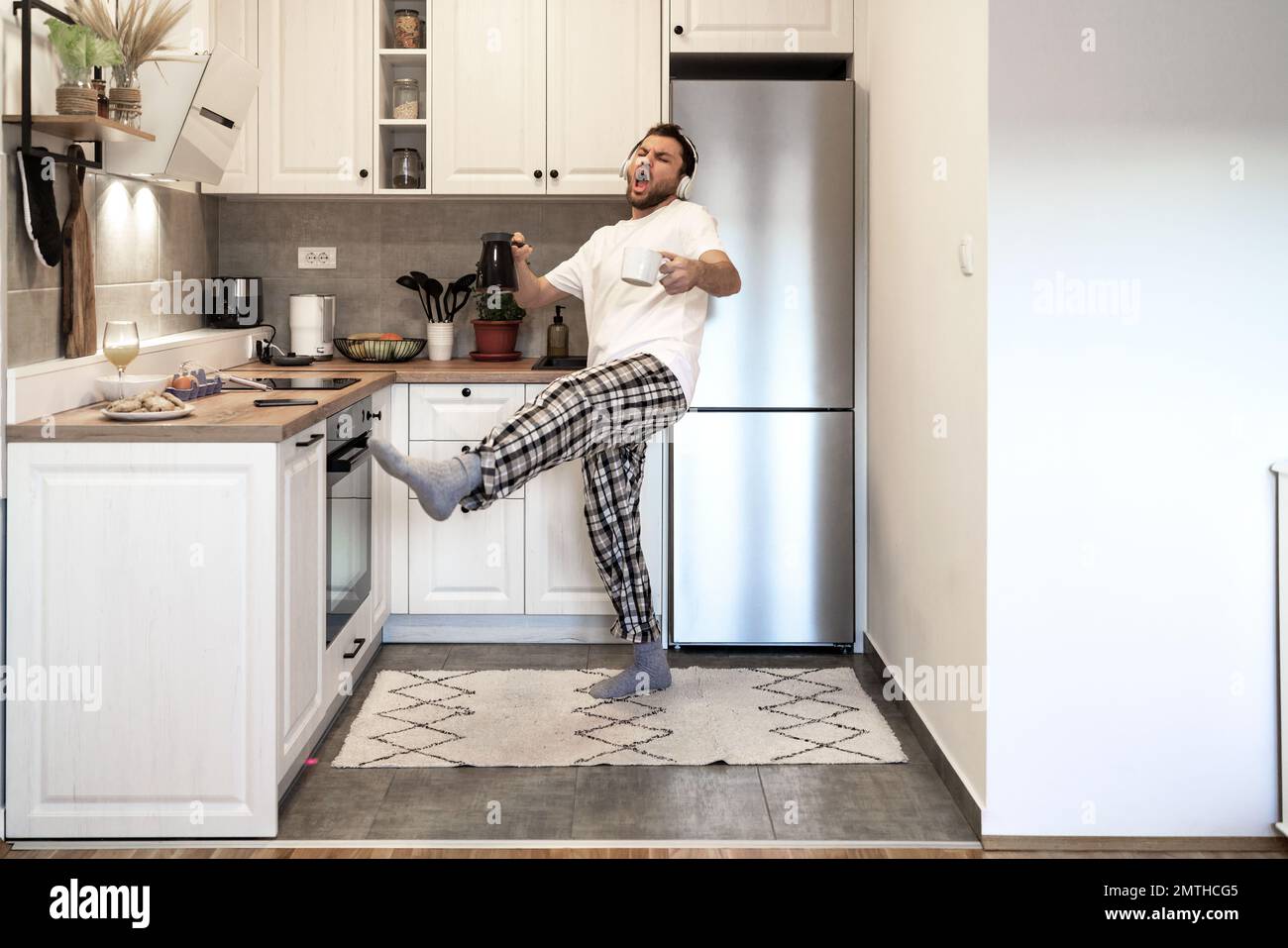 Ein bärtiger Mann in Pyjamahose und ein weißes T-Shirt tanzt und singt morgens mit Kopfhörern in der Hausküche, während er Kaffee kocht. Stockfoto