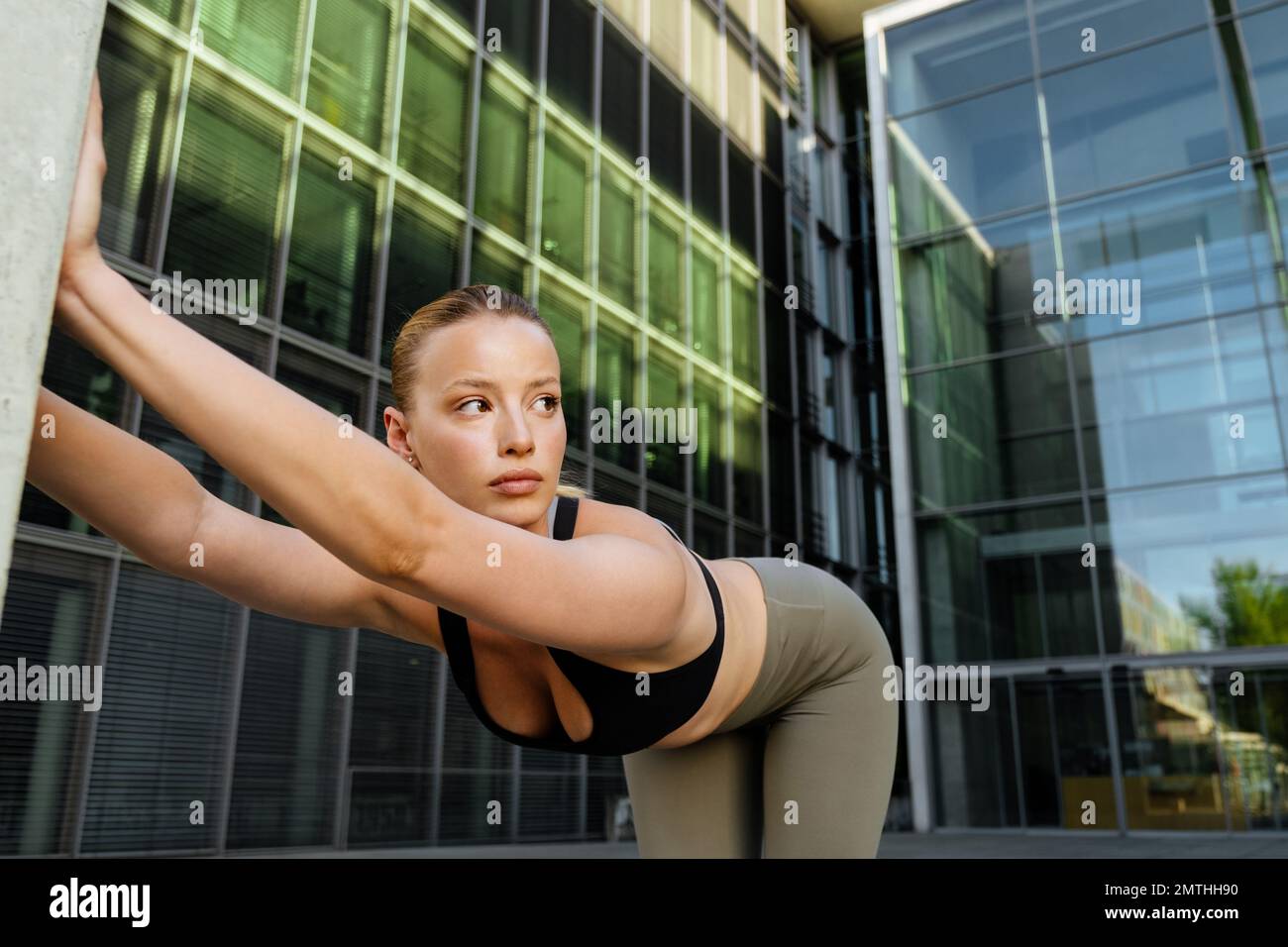 Junge, wunderschöne sportliche Frau, die sich den Rücken ausstreckt und zur Seite schaut, während sie neben einem modernen Gebäude steht Stockfoto