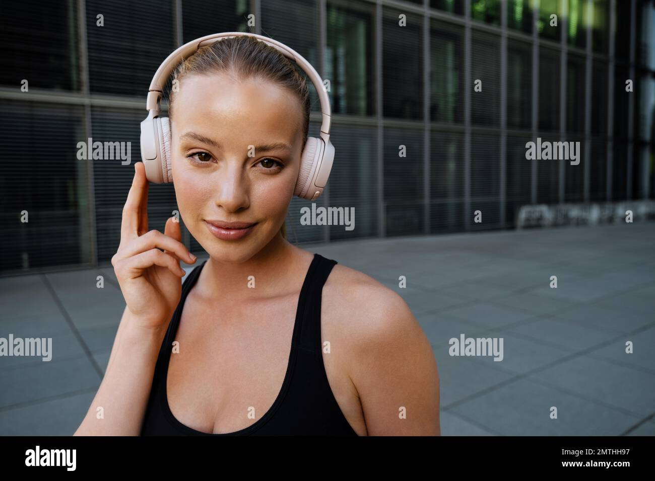 Junge schöne, attraktive, ruhige Frau, die Kopfhörer berührt und in die Kamera schaut, während sie auf der Straße neben einem modernen Gebäude steht Stockfoto