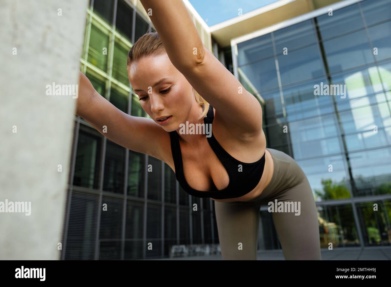 Junge, wunderschöne, sportliche Frau, die sich den Rücken ausstreckt und nach unten blickt, während sie in der Nähe eines modernen Gebäudes steht Stockfoto