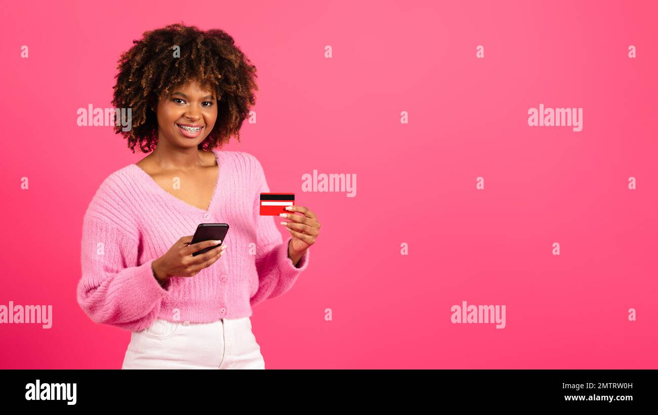 Die fröhliche junge schwarze, lockige Frau, Shopaholic in lässiger Klammer, benutzt Smartphone, Kreditkarte, um das Finanzkonto zu prüfen Stockfoto