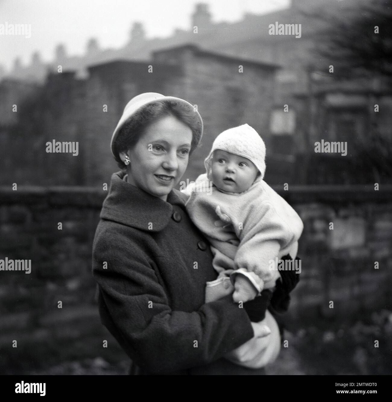 1950er, historisch, draußen in einem Garten eines terrassenförmigen Hauses, eine gut gekleidete Mutter mit Mantel und Hut, die für ein Foto steht, auf dem ihr Baby nach seiner Taufe gehalten wird, Northumberland, England, Großbritannien. Stockfoto