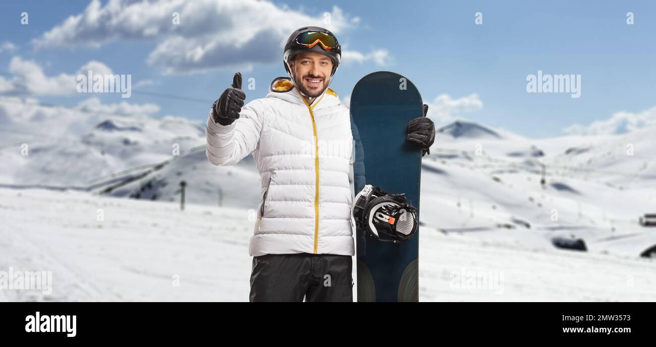 Glücklicher Mann mit Snowboardausrüstung, der auf einem Berg steht und Daumen nach oben zeigt Stockfoto