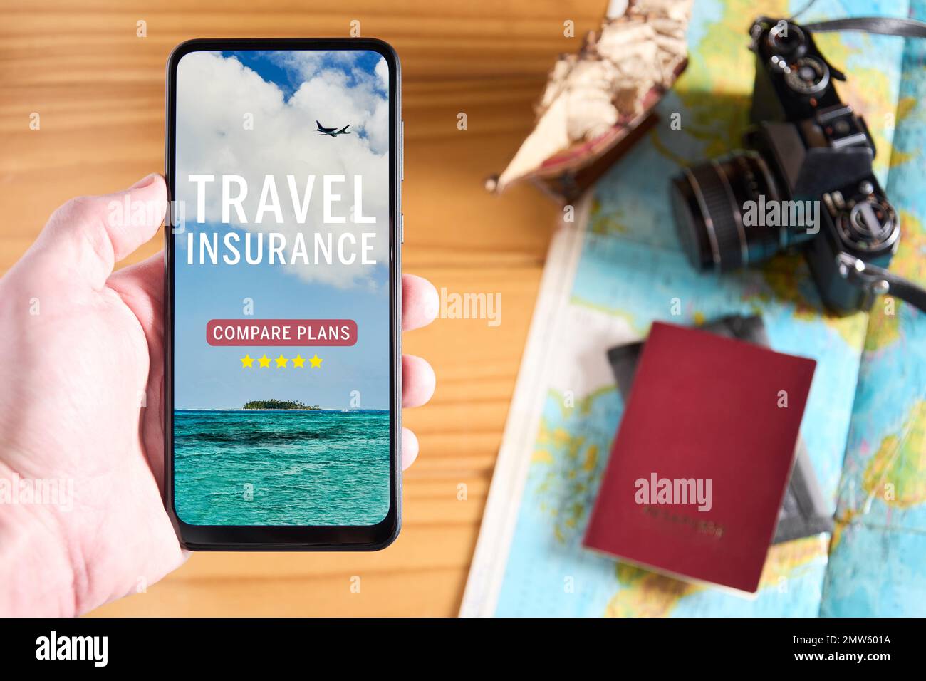 Draufsicht des Handys mit Reiseversicherungs-Website auf dem Bildschirm. Reisegegenstände auf Holztisch im Hintergrund mit Kamera, Pa Stockfoto