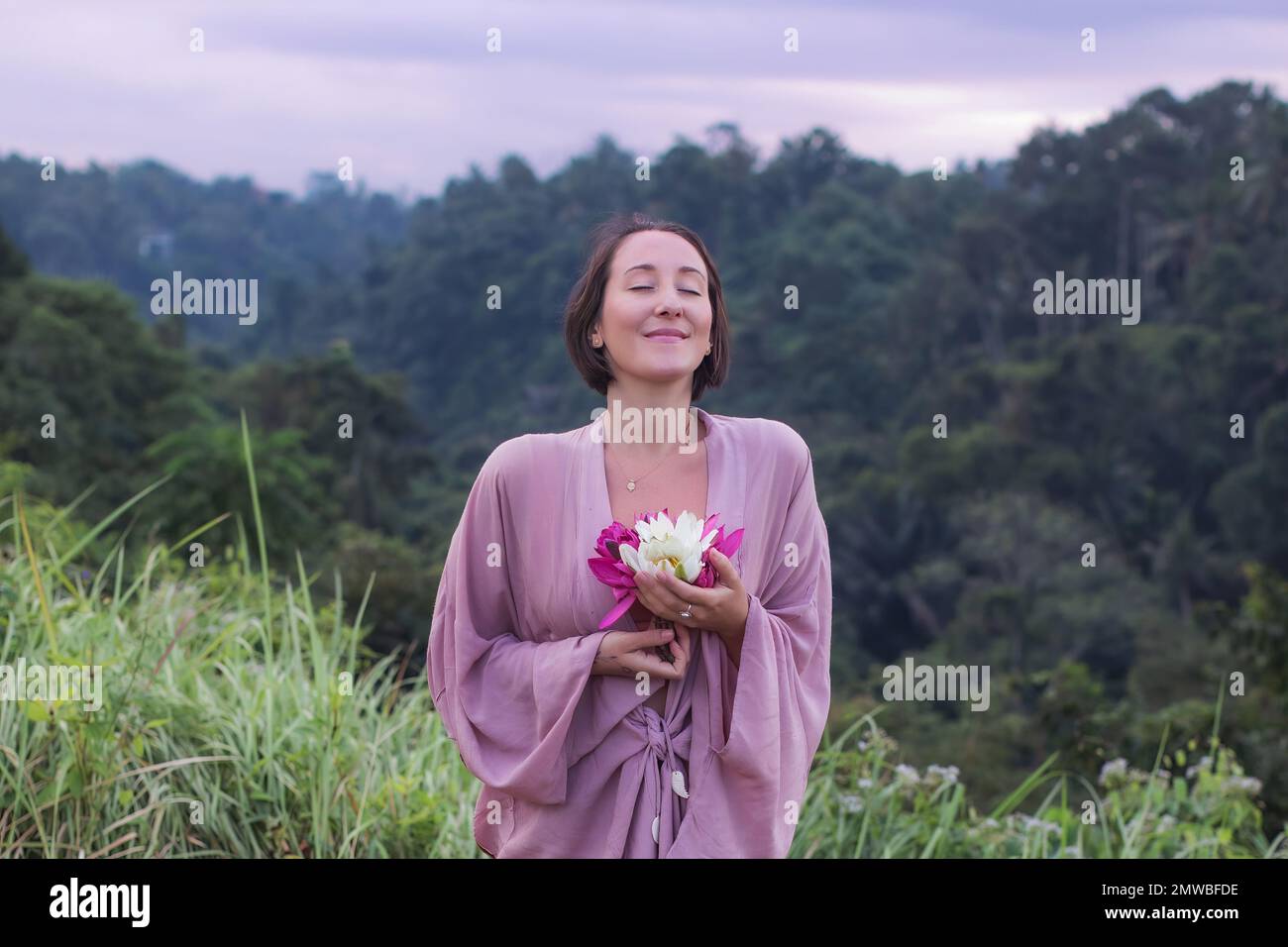 Eine Frau in einem violetten Bademantel, die am frischen Morgen in die Natur eintaucht und lila und weiße Blumen hält Stockfoto