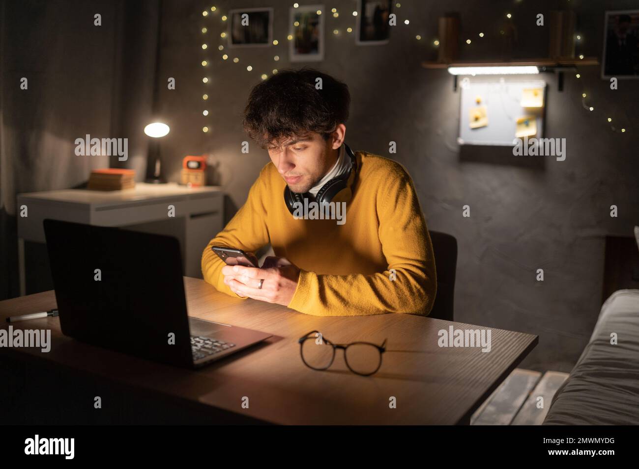 Ein junger Student, der im Studentenwohnheim studiert, am Schreibtisch sitzt, einen Laptop benutzt, ein Handy in der Hand hält. Speicherplatz kopieren Stockfoto