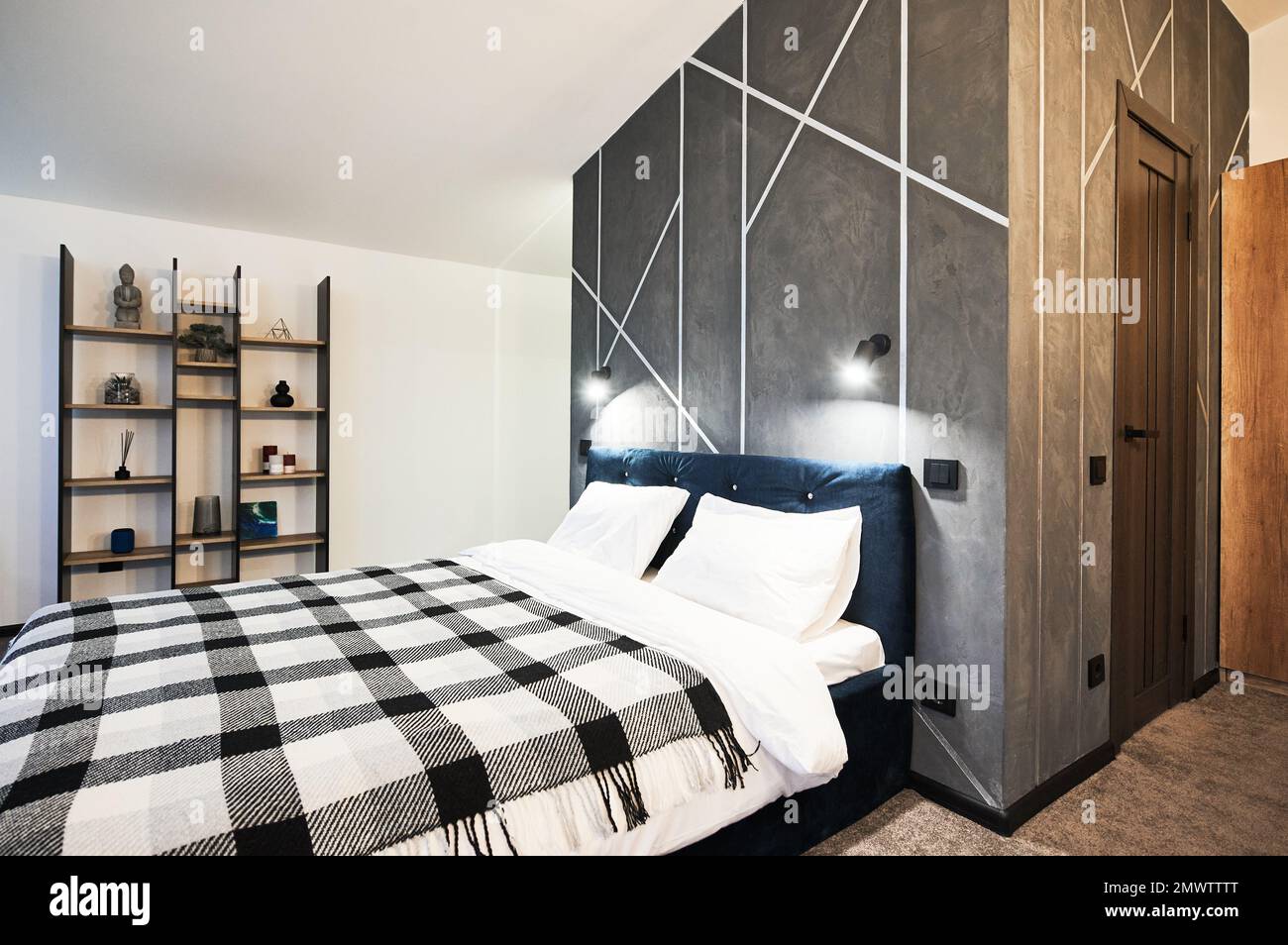 Schlafzimmer mit großem Bett und dekorierten Holzregalen im Innenbereich. Seitenansicht des breiten Bettes, mit kariertem Karomuster an grauen Wänden und geometrischem Muster im Schlafzimmer. Innenkonzept. Stockfoto
