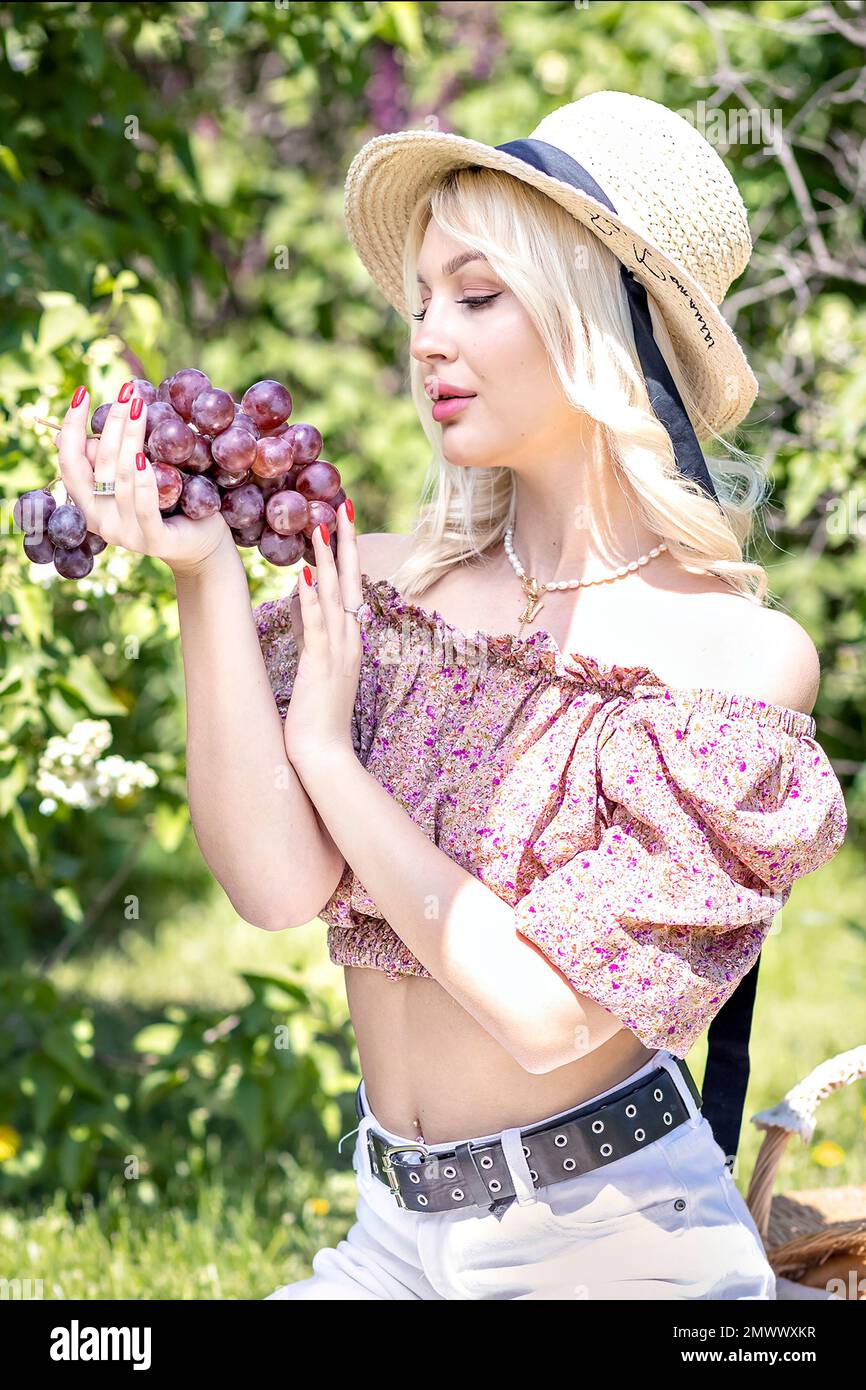 Foto einer jungen blonden Frau mit Hut, die einen Haufen Trauben in der Hand hält. Erholung im Freien, Picknick im Sommer. Stockfoto