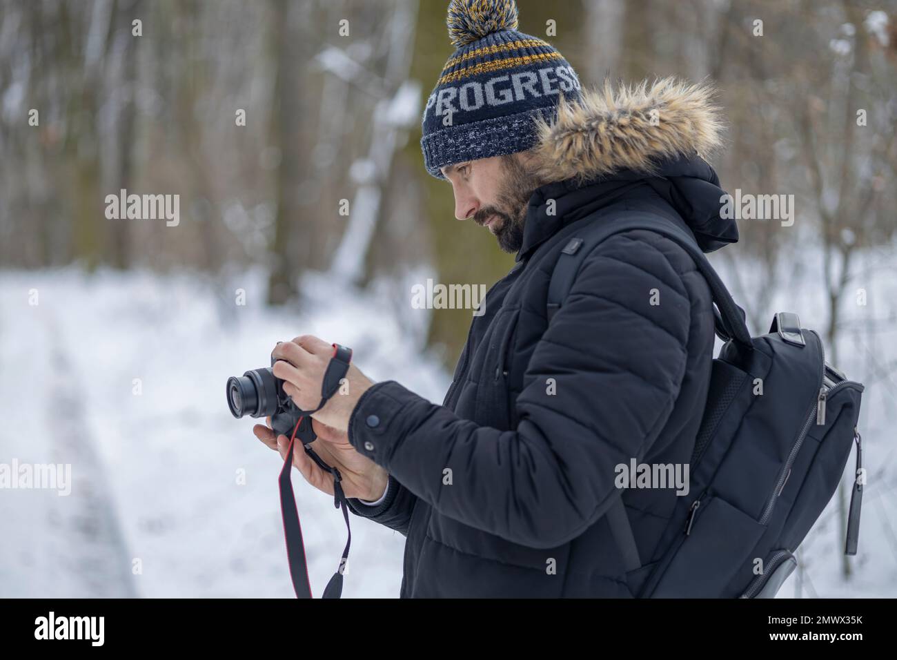 Ein Mann mit einem Bart im Wald im Winter hält seine Kamera und überprüft etwas auf dem camera.in verschwommenen Hintergrund, es gibt Wald und Schnee Stockfoto