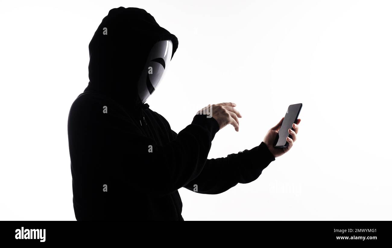 Anonymer Hacker und Gesichtsmaske mit Smartphone in der Hand. Mann in schwarzem Kapuzenhemd, der ein Mobiltelefon auf weißem Hintergrund hält und benutzt. Repräsentiere Cyber cr Stockfoto