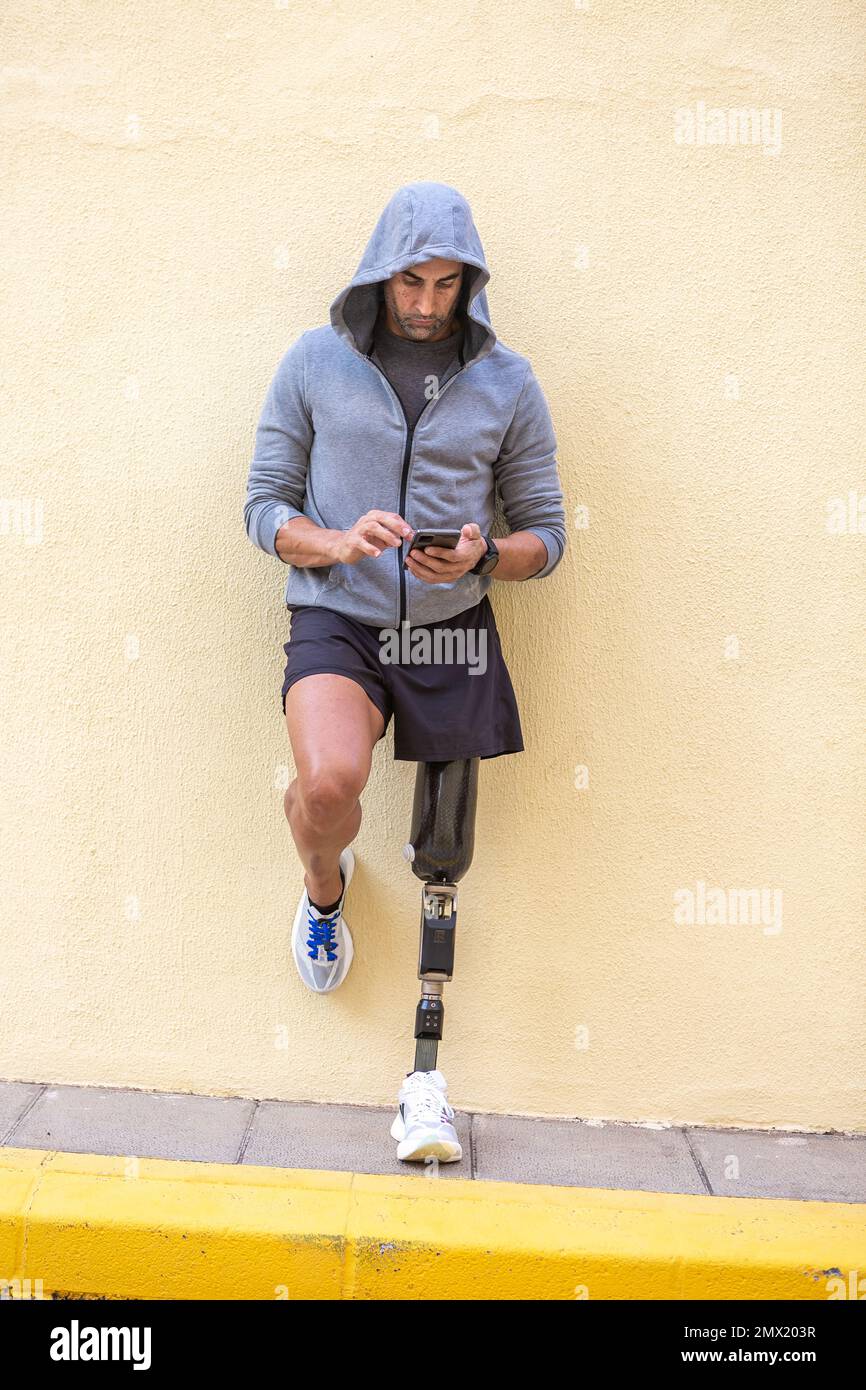 Ganzkörpersportler mit Beinprothese lehnt sich an die gelbe Wand und surft im Handy, während er eine Pause beim Fitnesstraining auf der Straße macht Stockfoto