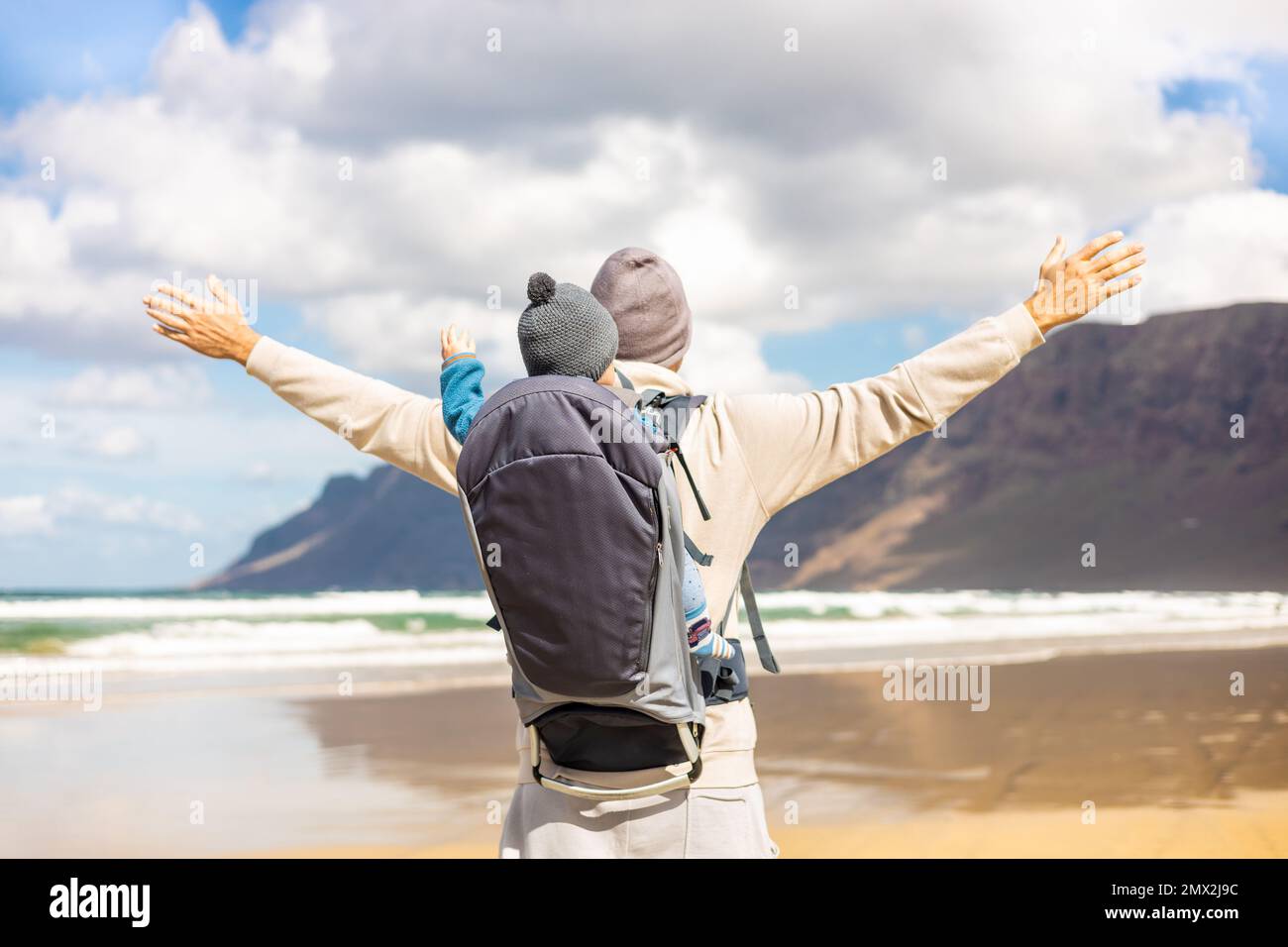 Der junge Vater erhebt sich die Hände zum Himmel und genießt dabei die reine Natur, während er seinen kleinen Sohn im Rucksack am windigen Sandstrand von Famara, Lanzarote, trägt Stockfoto
