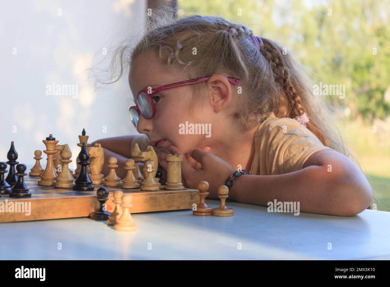Ein kleines Mädchen mit langen blonden Haaren und Brille spielt Schach Stockfoto