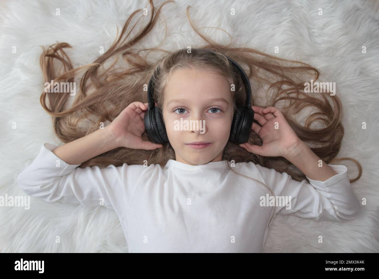 Kleines Mädchen mit kabellosen Kopfhörern auf einem weißen Pelzteppich Stockfoto
