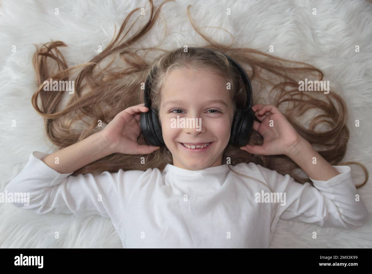 Kleines Mädchen mit kabellosen Kopfhörern auf einem weißen Pelzteppich Stockfoto