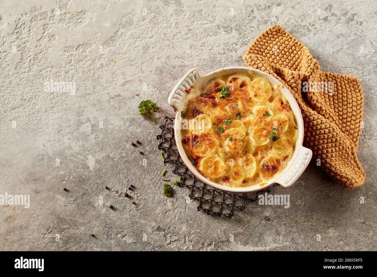 Draufsicht auf schmackhafte, kallopierte Gratin-Kartoffel mit Kräutern und Metallständer für heißes Gericht in der Nähe des Handtuchs auf grauer Steinoberfläche Stockfoto