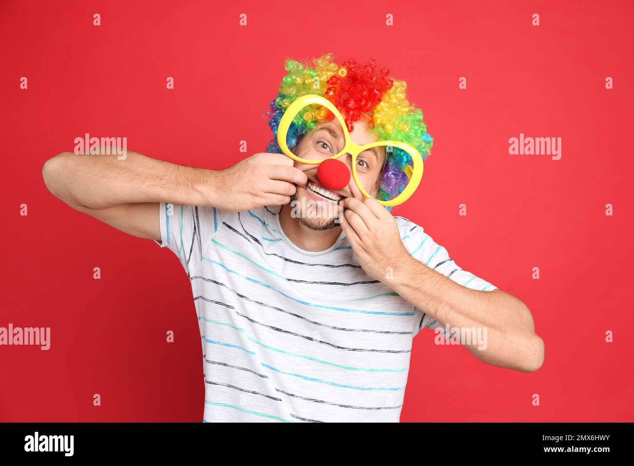 Lustiger Mann mit großer Brille, Regenbogenperücke und Clownnnase auf rotem Hintergrund. Der Aprilscherztag Stockfoto
