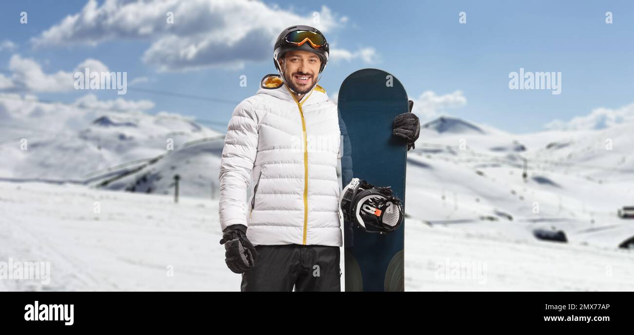 Ein Mann mit Schutzbrille und Helm, der ein Snowboard auf einem Berg hält und vor der Kamera lächelt Stockfoto