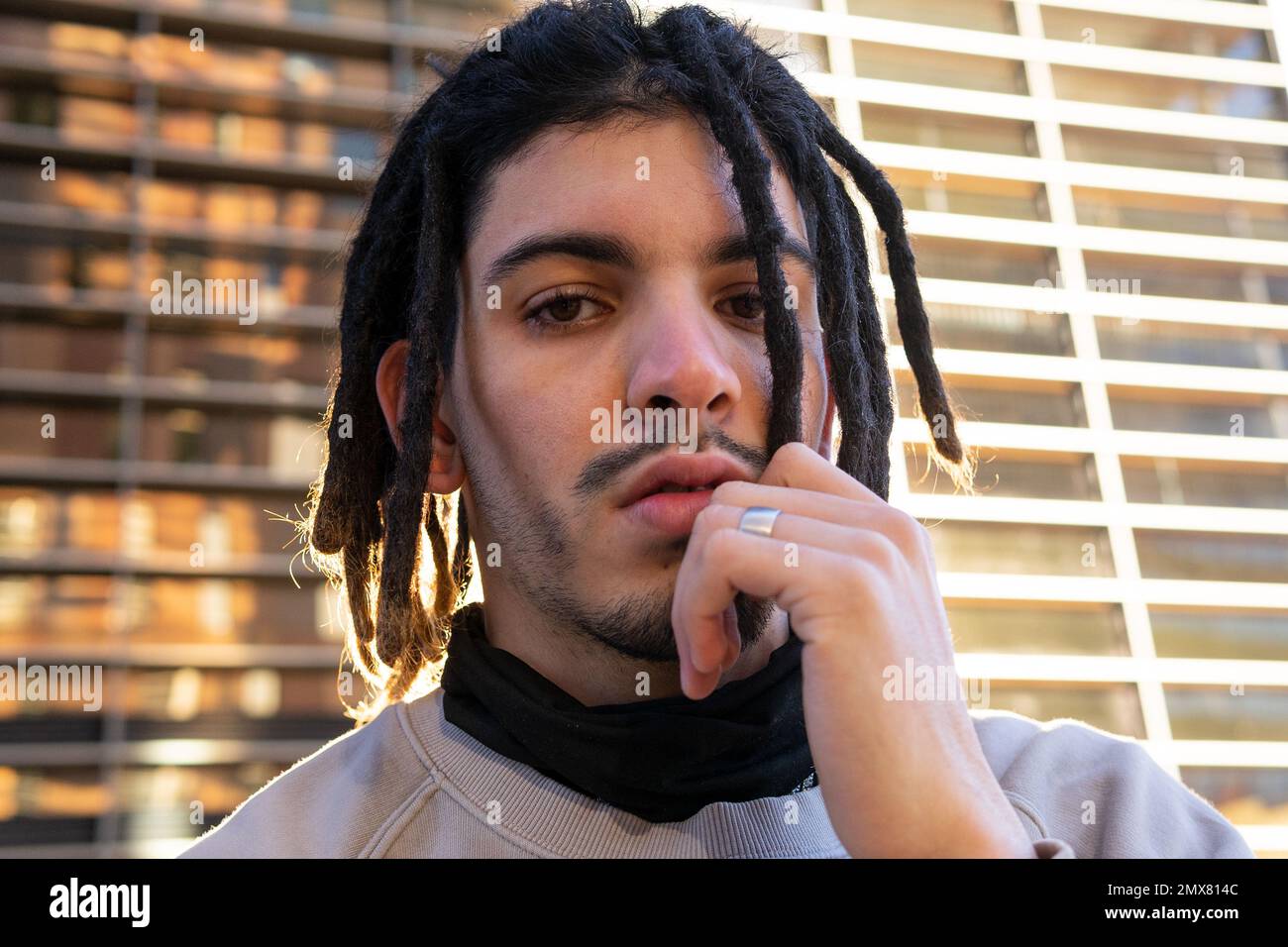 Kleiner Winkel eines nachdenklichen jungen Mannes mit Dreadlocks, der in die Kamera schaut, während er sein Gesicht berührt und gegen ein modernes Gebäude steht Stockfoto