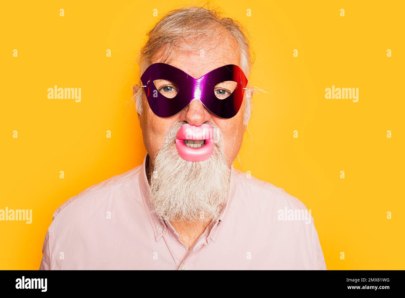 Positiv bärtiger alter Mann in rosa Hemd trägt Karnevalsmaske und lustige Lippen, die auf gelben Hintergrund auf die Kamera schauen Stockfoto