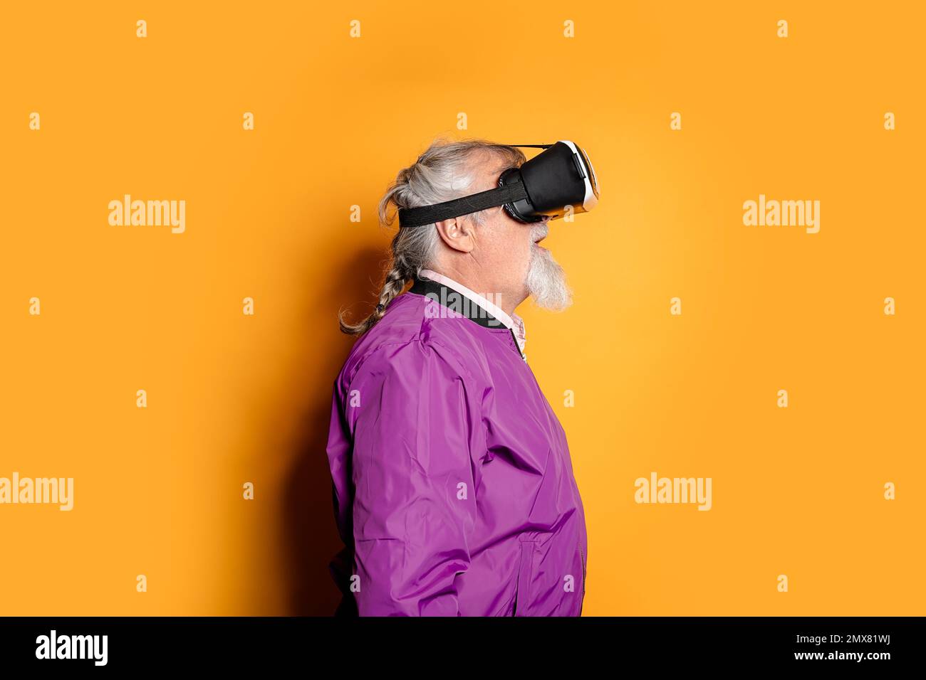 Porträt eines bärtigen älteren Mannes, der eine VR-Schutzbrille und eine lila Jacke trägt und vor orangefarbenem Hintergrund steht, während er im Studio den Cyberspace erkundet Stockfoto
