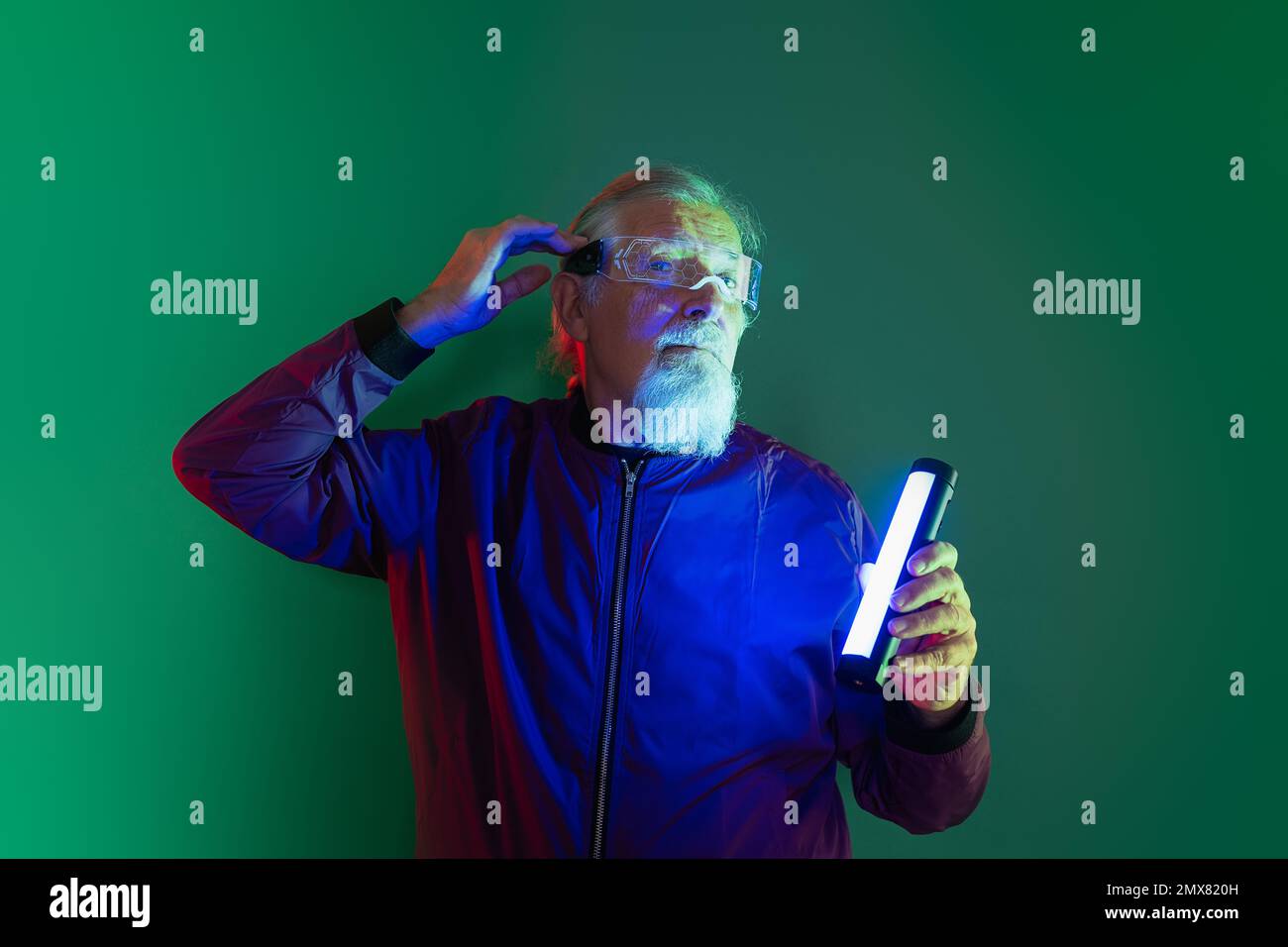 Überraschter männlicher Hipster mit Bart in Brille und lässiger Kleidung, die den Kopf berührt, während er im Studio vor grünem Hintergrund mit Neonlicht steht Stockfoto