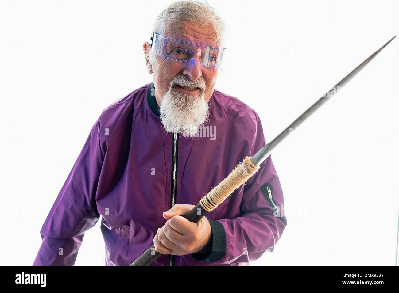 Lächelnder älterer bärtiger männlicher Hipster in Brille, der vor weißem Hintergrund mit scharfem Schwert steht Stockfoto