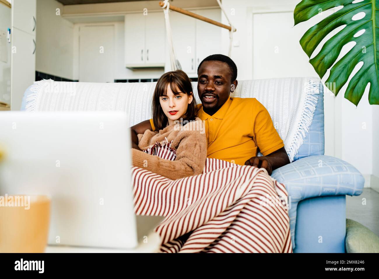 Ein junges, multiethnisches Paar in legerer Kleidung, das auf dem Sofa sitzt und auf dem Laptop surft, während es seine Freizeit zu Hause verbringt Stockfoto