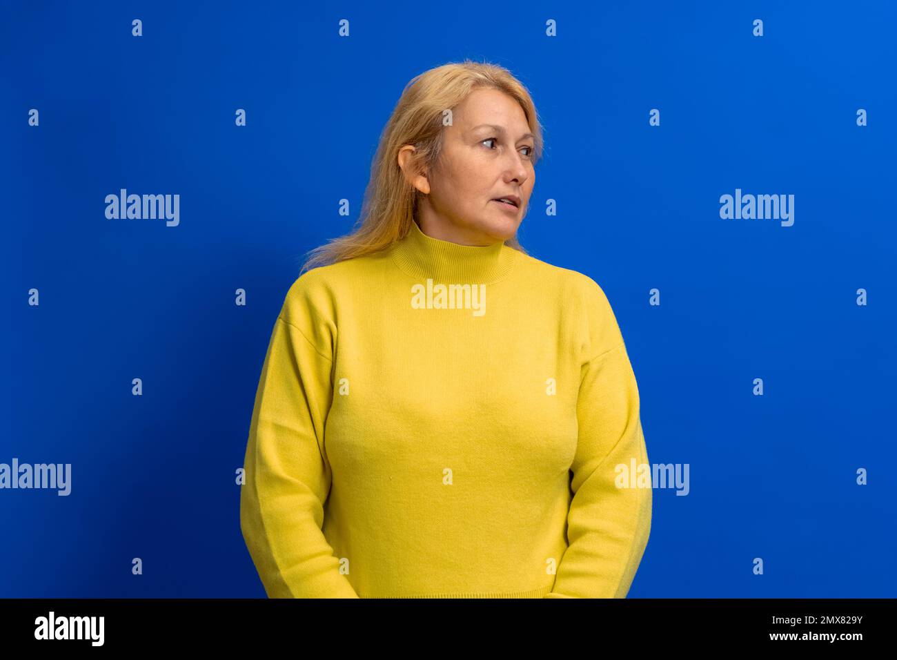 Wunderschöne Frau in ihren 40s Jahren, die einen gelben Pullover trägt, auf blauem Hintergrund steht und auf die Seite schaut, mit einem ernsthaften Gesicht. Stockfoto