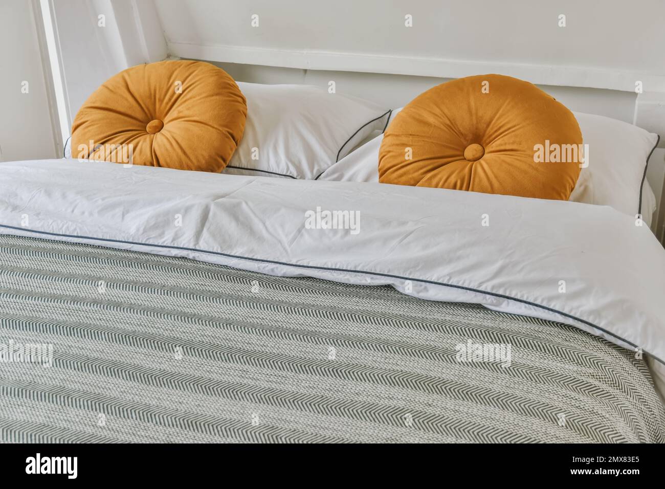 Weiches Bett mit Kissen und Decke, dekoriert mit orangefarbenen Kissen an der weißen Wand im hellen Schlafzimmer zu Hause Stockfoto