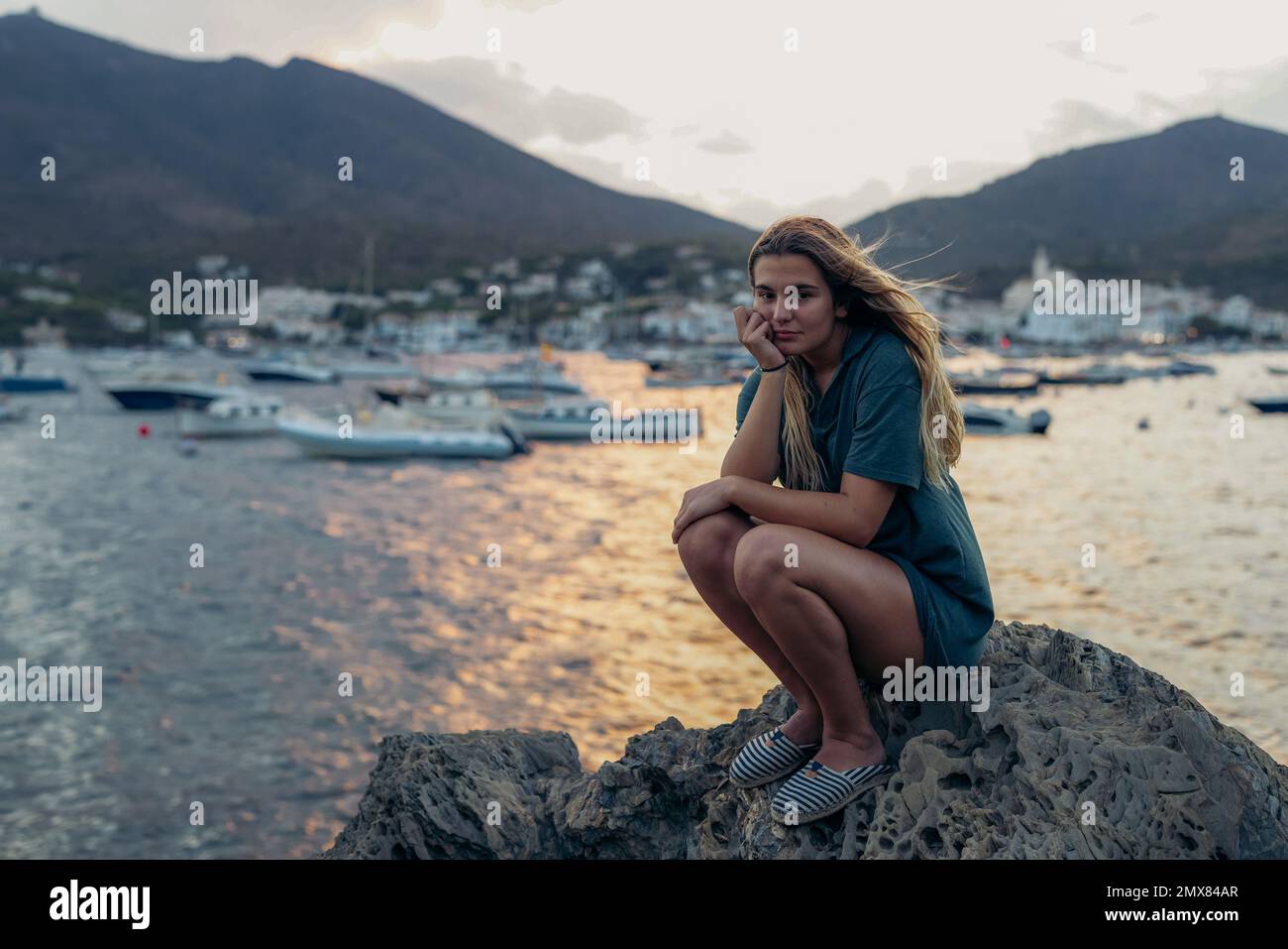 Voller Körper von jungen, ruhigen, aufmerksamen weiblichen Touristen mit langen blonden Haaren im T-Shirt, die auf einem Felsbrocken sitzen, mit der Hand am Kinn sitzen und auf Cadaqu in die Kamera schauen Stockfoto