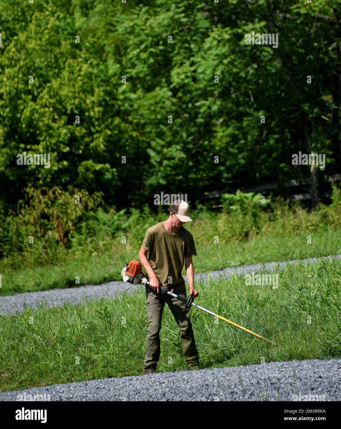 Ein junger Mann hält Unkrautfresser und schneidet das Gras über die Schottereinfahrt. Er ist verschwitzt und heiß, während er in der Sommerhitze arbeitet. Stockfoto