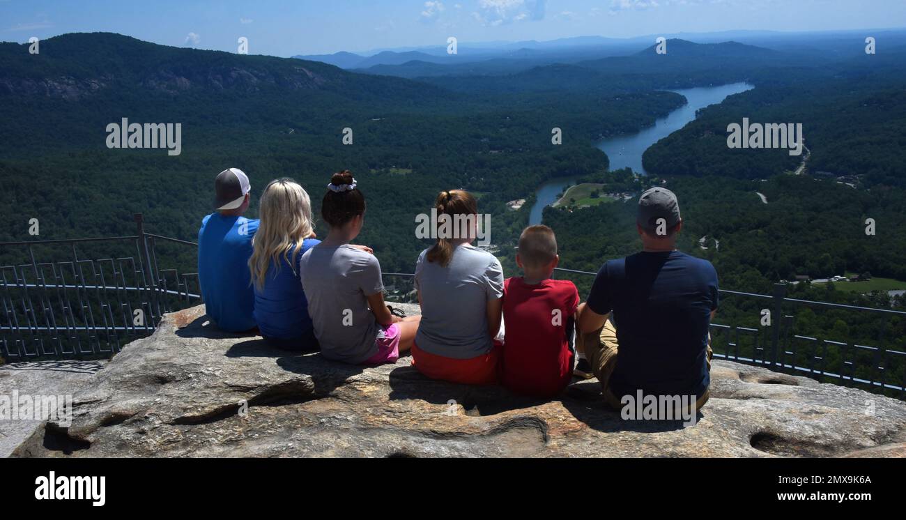 Eine sechsköpfige Familie sitzt am Rand des Felsens und bewundert die Aussicht vom Chimney Rock, Chimney Rock State Park, North Carolina. Seehund ist in der Ferne zu sehen. Stockfoto