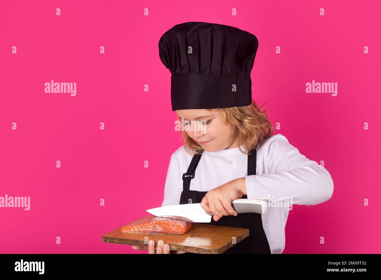 Kinderkoch hält Schneidebrett mit Lachssteak und Messer. Koch-Kind bereitet gesundes Essen zu. Porträt eines Kindes mit Koch-Hüten. Stockfoto