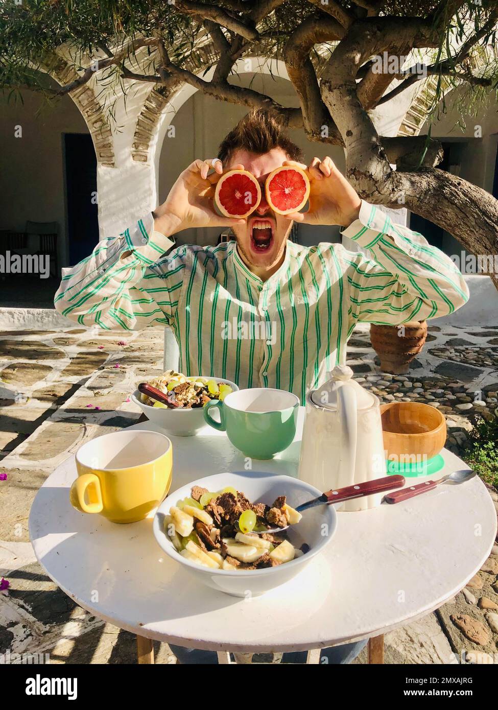 Junger Mann im Hemd, grimpert und hält eine halbe Grapefruit als Augen vor seinem Gesicht, Frühstück mit Müsli und Tee auf einer Terrasse, Paros Stockfoto