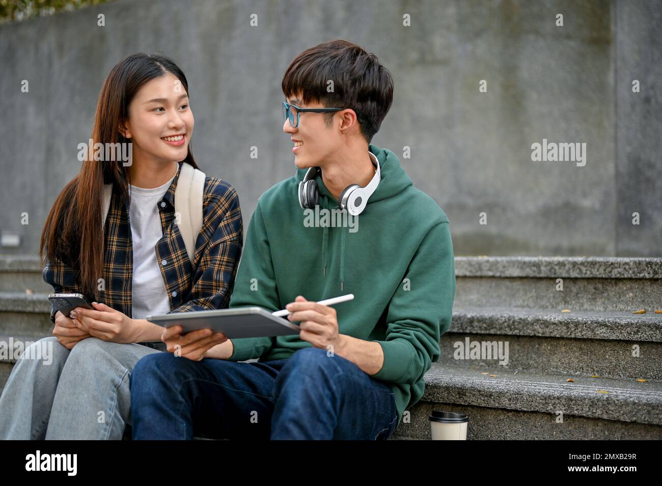 Zwei glückliche junge asiatische College-Freunde, die auf der Treppe sitzen und ein Tablet benutzen, unterhalten sich gern und diskutieren über ihre Schulprojekte. Stockfoto