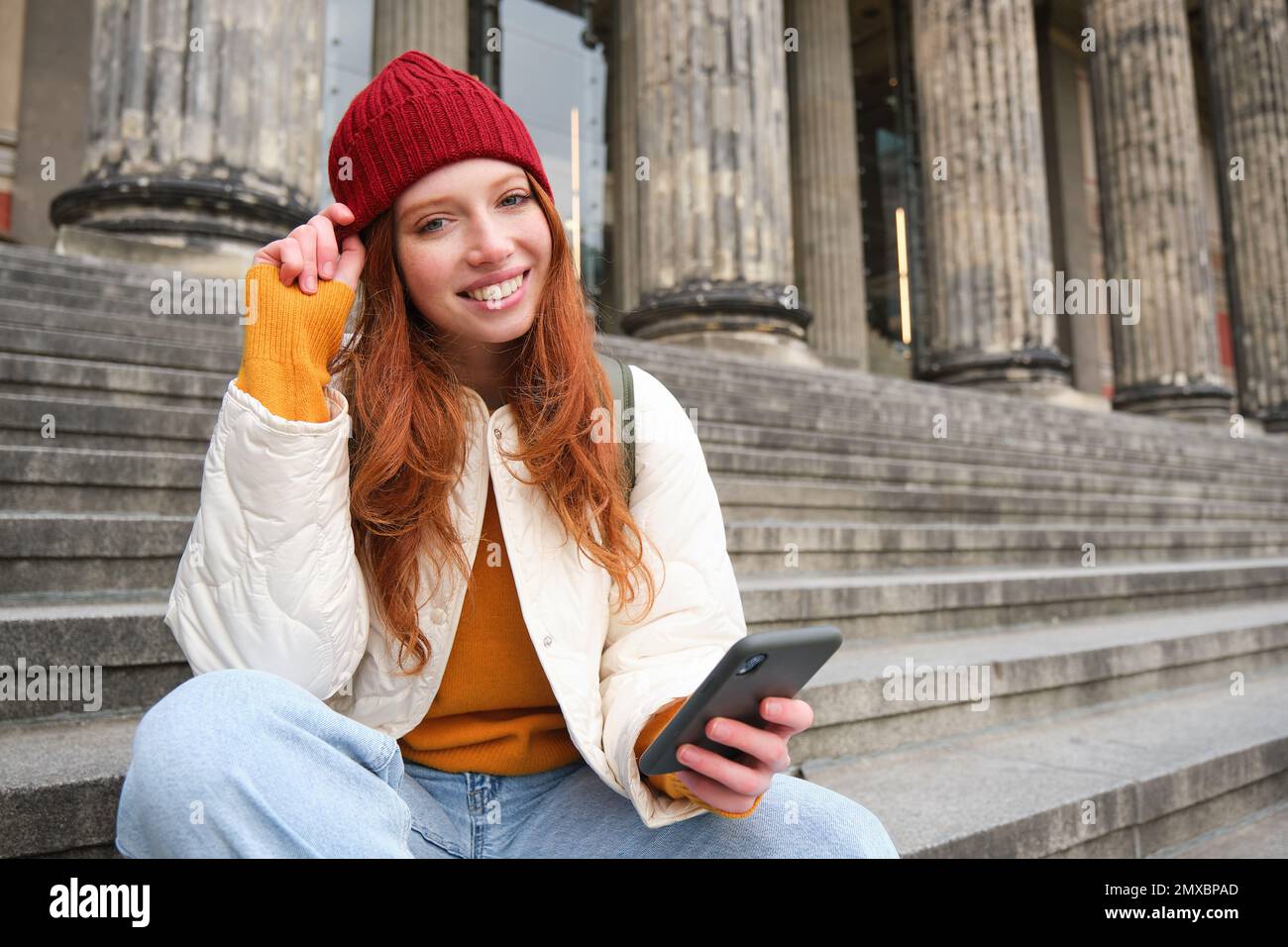 Porträt eines jungen Stadtmädchens mit rotem Hut, sitzt auf der Treppe in der Nähe des Museums, hält ein Mobiltelefon, stellt eine Verbindung zu öffentlichem WLAN und Surfnetz her, verwendet Smartphone-Apps. Stockfoto