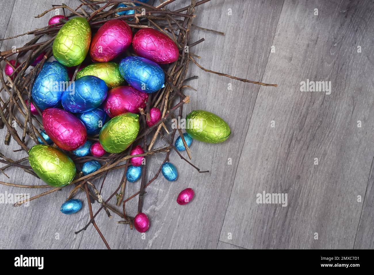 In Folie gewickelte mehrfarbige ostereier in rosa, grün, blau und gelb in einem natürlichen Nest aus Stäben und Zweigen, gegen einen grau weißen Holzhinterholz Stockfoto