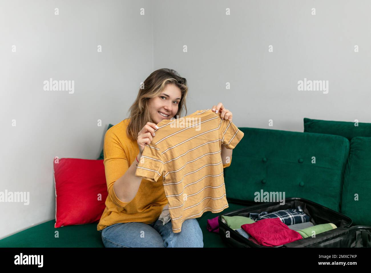 Lächelnde Frau, gestreiftes T-Shirt, das auf dem Sofa im Wohnzimmer sitzt. Glückliche Frau faltete Freizeitkleidung in ihr Gepäck. Stockfoto