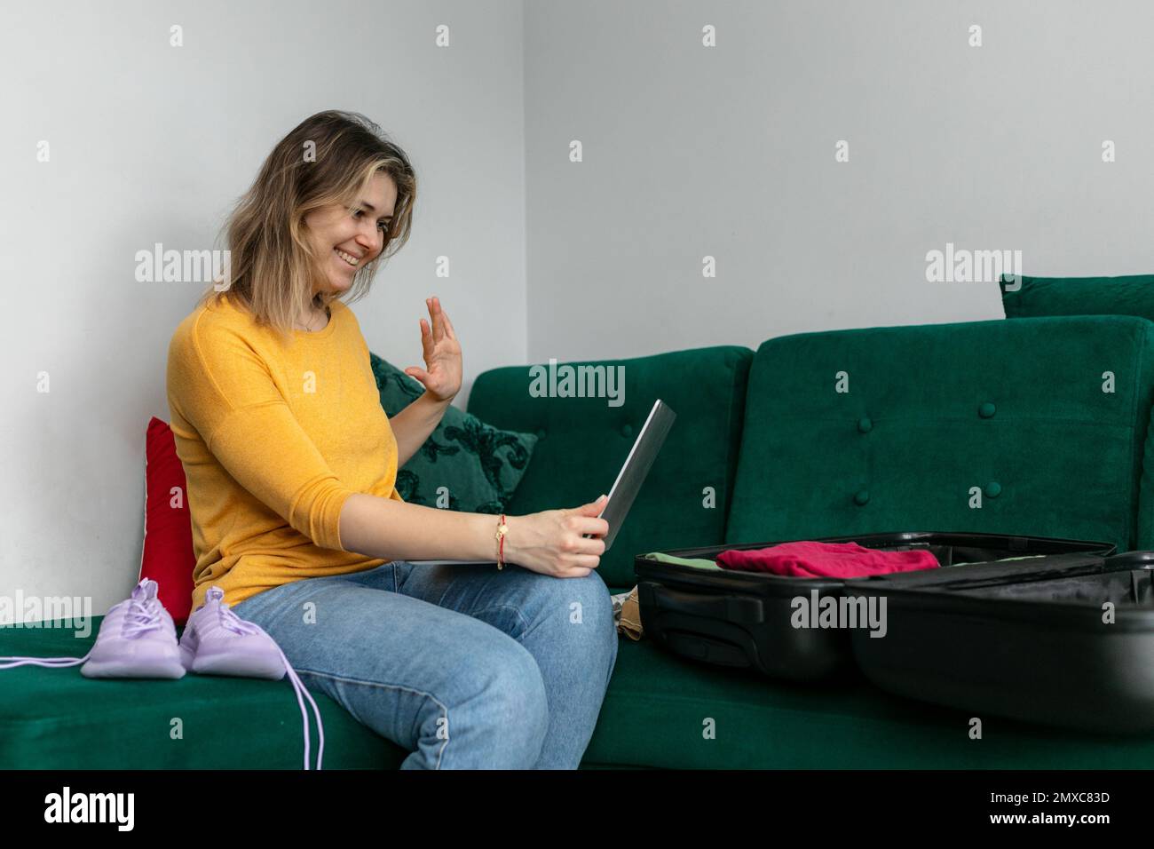Seitenansicht einer lächelnden jungen Frau, die auf einem grünen Sofa sitzt, auf den Bildschirm des Tablets schaut, mit der Hand winkt, Videoanrufe führt. Stockfoto