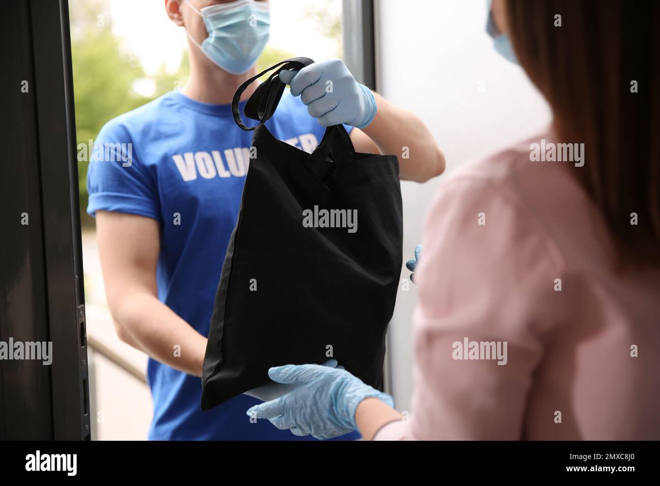 Männlicher Freiwilliger gibt der Frau schwarze Tasche durch die Tür, schließen. Hilfe während der Coronavirus-Quarantäne Stockfoto