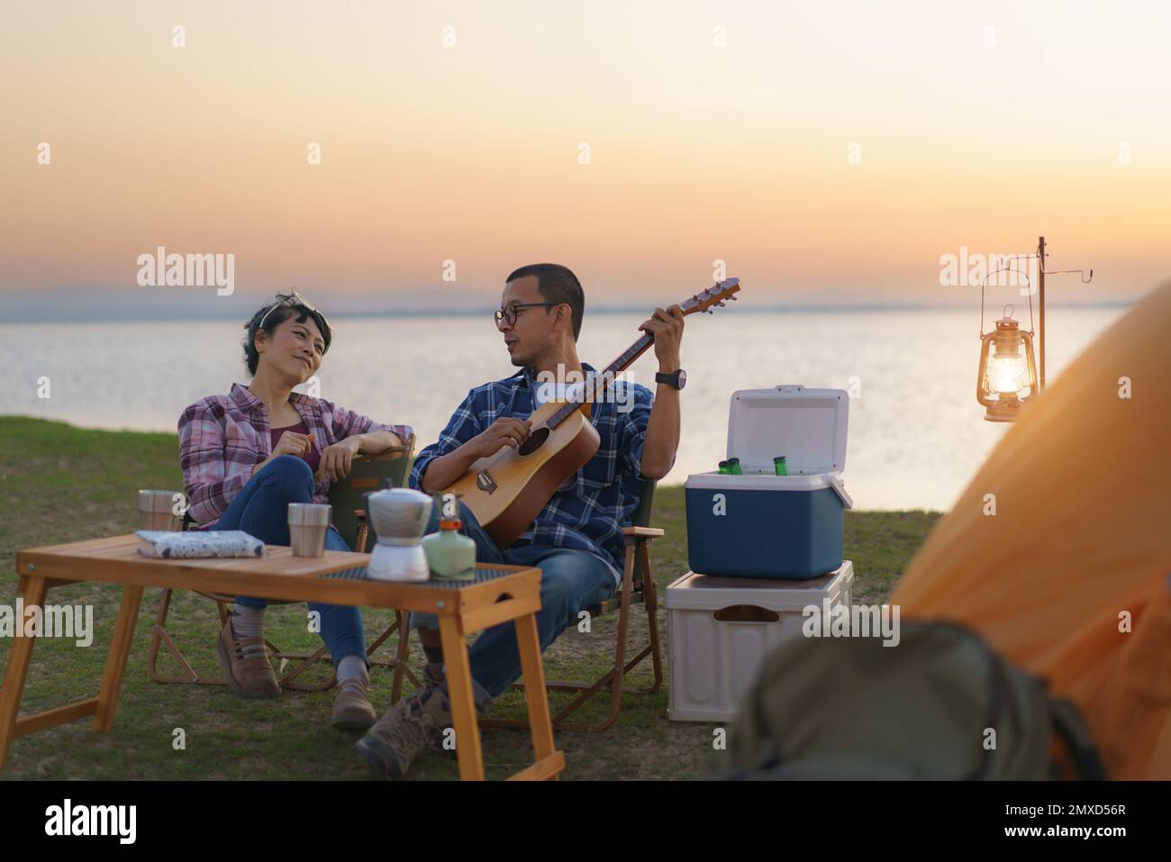 Ein asiatisches Paar singt und spielt Gitarre in seinem Campingbereich mit See im Hintergrund während des Sonnenuntergangs. Stockfoto